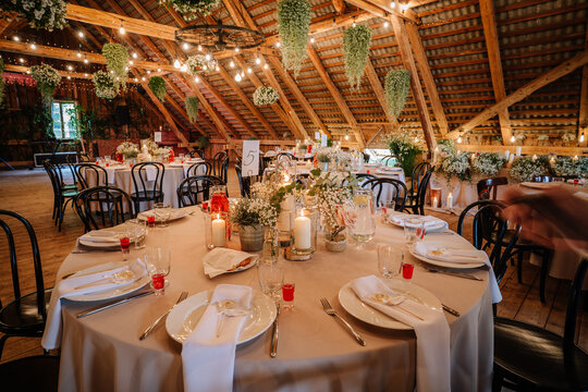 Rustic wedding reception setup with round tables, floral centerpieces, candles, string lights, hanging greenery, and elegant table settings in a wooden hall.