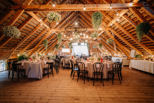 Beautiful rustic wedding reception hall with wooden beams, round tables, floral centerpieces, candles, string lights, and hanging greenery in a cozy venue.