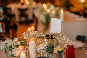 Close-up of an elegant wedding table centerpiece with candles, flowers, greenery, a blank place card, lemon water, and red drink in a cozy rustic venue.