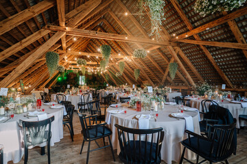 Rustic wedding reception setup in a wooden barn attic, decorated with fairy lights, floral arrangements, candles, and round tables covered in beige cloths.