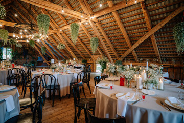 Rustic barn wedding reception with round tables, floral centerpieces, candles, and string lights, set under a charming wooden-beamed attic ceiling.