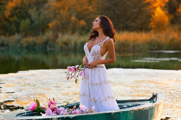 Beautiful woman in white dress on a boat with flowers on a lake in the evening light.