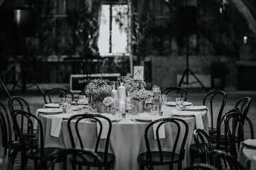 Black and white photo of an elegant round wedding table with candles, flowers, and number card, set in a rustic indoor venue with wooden chairs.