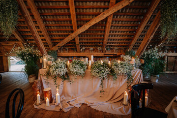 Beautifully decorated sweetheart table in a rustic venue with floral arrangements, candles, logs, and hanging greenery, perfect for a romantic wedding celebration.