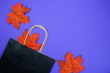 Black shopping bag with autumn maple leaves on a purple background. Concept of seasonal sales, online shopping, gifts for Halloween, Thanksgiving day, back to school and autumn discounts. Copy space. 