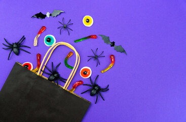 Black paper bag with holiday candies, gummy worms, toy spiders, bats and eyeballs on purple background. Halloween shopping and celebration concept. Top view. Copy space.