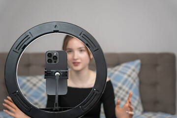 Young girl vlogger recording video with smartphone using ring light while sitting on bed at home, creating engaging content for social media platforms. Focus on the ring lamp