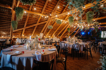 A beautifully decorated rustic wedding reception hall with round tables, floral centerpieces, candles, wooden beams, and hanging greenery under warm lighting.