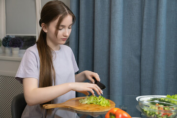 Young girl is carefully chopping fresh lettuce on a wooden cutting board, preparing a healthy salad with other vegetables like tomatoes, cucumbers, and dill in her modern kitchen