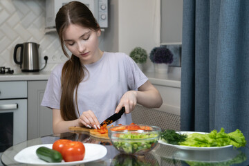 Young girl is carefully chopping fresh vegetables on wooden cutting board, preparing a healthy meal or salad in her modern kitchen, surrounded by various ingredients