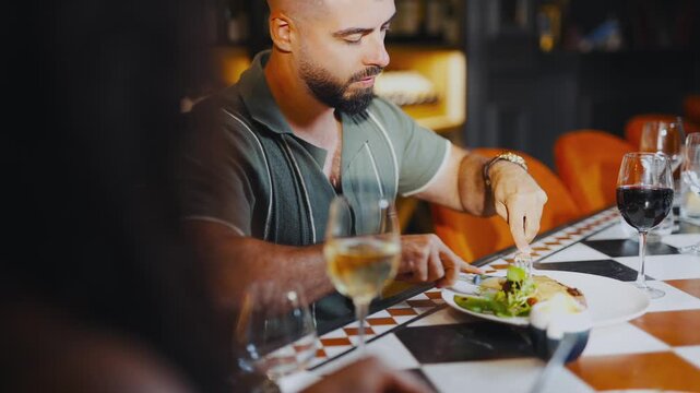 A man is eating at a restaurant with a woman in the background. He is using a knife and fork to cut his food