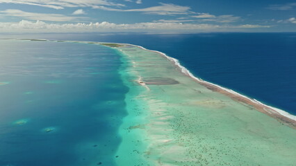 Crystal clear turquoise waters stretch out around a narrow sandy islet under a sunny sky. The coastline features stunning coral reefs that teem with aquatic life.