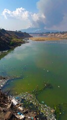 A polluted river winds through a landscape, choked with trash and algae, under a partly cloudy sky.