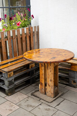 Round wooden table and benches made from recycled pallets in outdoor seating area near white wall and window with flowers.