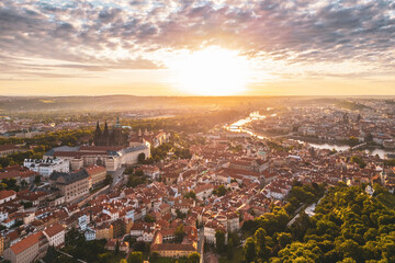 Sunrise casts a warm glow over Prague Castle and the vibrant rooftops of the city. The picturesque landscape, with rivers and greenery, captures a serene morning atmosphere in Praha.