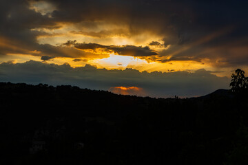 A stunning sunrise over a dark, silhouetted hill landscape with sunrays breaking through thick, dramatic storm clouds creating a powerful and beautiful contrast.
