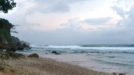 Waves gently roll onto a serene rocky beach at dusk, with lush green cliffs on either side under a dramatic, cloudy sky.
