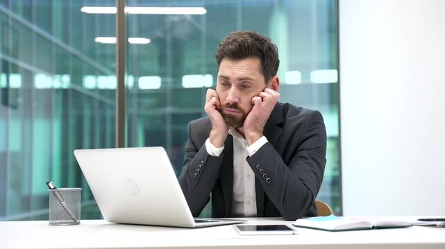 Overworked businessman is bored sitting at workplace at desk in business office. Exhausted employee in formal suit is tired from working with laptop. A lazy male worker procrastinates, gets distracted