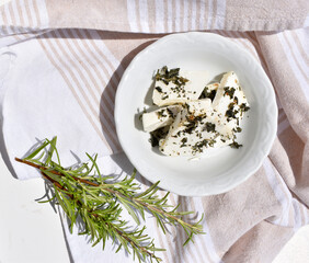 Fresh cottage cheese slices served on a white plate and decorated with rosemary sprig. Rustic farm style healthy vegetarian snack, top view, flat lay food photography.