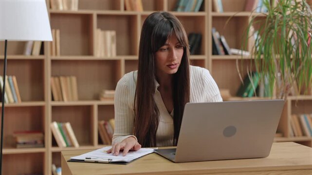 Serious beautiful Caucasian woman leaning forward and analyzing printed data while touching report. Preparing presentation or double-checking numbers before sharing results in virtual meeting.