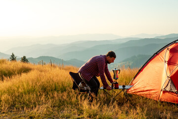 Young man on campsite sets up a table with coffee near a red tent with mountain peaks in the background at sunset in summer in Pamporovo, Bulgaria. Active vacation with family. Nature background