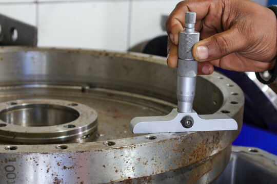 A close-up of a person's hand using a depth micrometer to precisely measure a large industrial metal component. The focus is on the intricate process of quality control and precision.