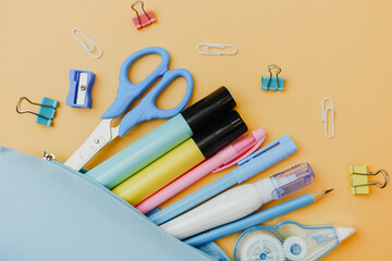 A collection of colorful pens, scissors, and office supplies spilled from a light blue pouch onto a soft orange background.