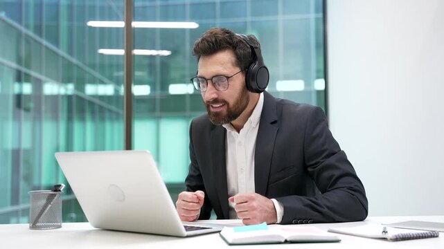 Excited joyful businessman in wireless headphones watching sports match or competition using laptop sitting at workplace in business office. Happy man cheering for bids at auction, celebrating success