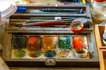 Painting set with watercolor paints, brushes, and palette on wooden table, used in artist’s studio for creating traditional artworks.