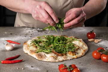 Hands of a chef adding fresh arugula to a delicious pizza topped with cheese, garlic, and cherry tomatoes, showcasing culinary artistry and vibrant ingredients
