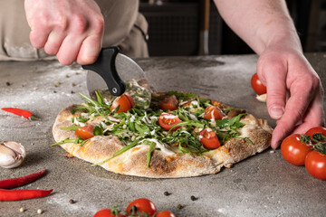 Chef using a pizza cutter to slice a freshly baked pizza topped with tomatoes, arugula, and drizzled sauce on a rustic kitchen countertop, showcasing culinary skills and delicious food