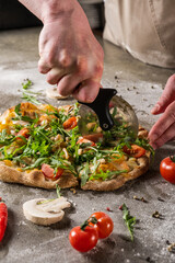 Hands of a chef using a pizza cutter to slice a freshly baked pizza topped with arugula, tomatoes,...