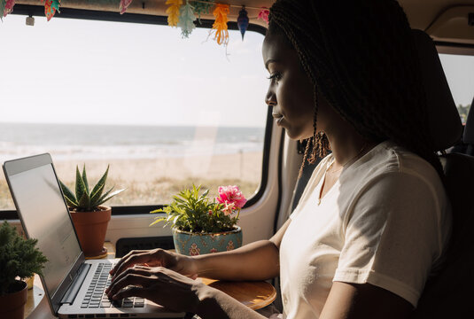 focused woman working remotely on laptop in a cozy van with ocean view, surrounded by potted plants - Powered by Adobe