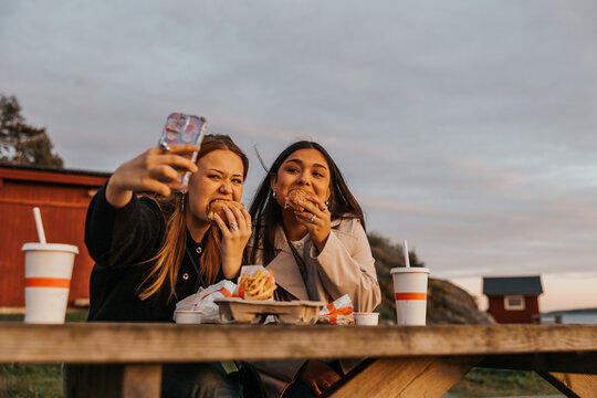 Young woman taking selfie with friend while eating burger at picnic table - Powered by Adobe