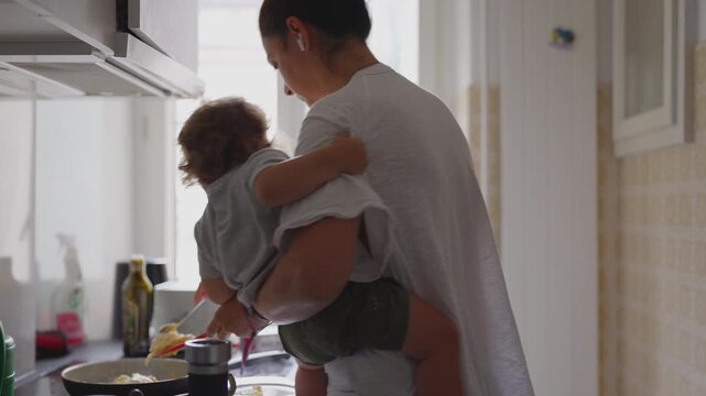 Mother cooking in kitchen while holding toddler in her arms, multitasking between preparing food and comforting child during candid authentic family moment at home