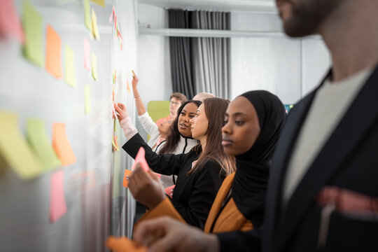 Female business experts discussing ideas while sticking adhesive notes on whiteboard with colleagues in office meeting room