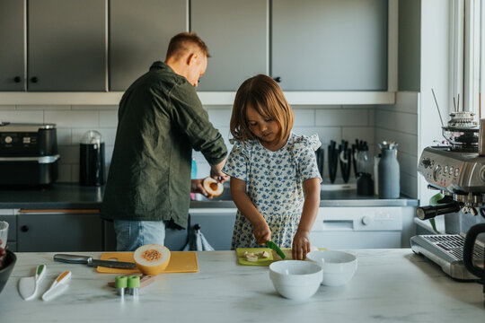 Cute girl cutting banana while father cleaning melon in background at kitchen