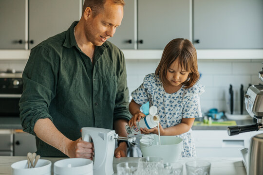 Father assisting daughter pouring milk in bowl while standing in kitchen at home