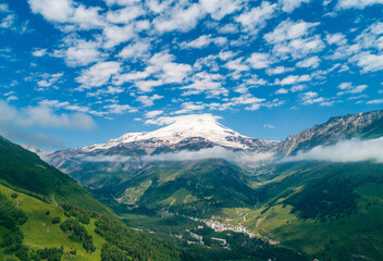 Majestic view of Mount Elbrus towering over lush green valleys under a bright blue sky with fluffy clouds