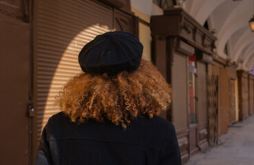 Authentic urban portrait of woman with curly hair and black beret in Saint Petersburg, back view. Warm clothes, historic arches, lanterns and cultural atmosphere reflect city lifestyle in Russia
