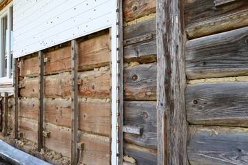 A part of an old wooden house's facade where the cladding has been dismantled, revealing weathered logs and remnants of insulation.