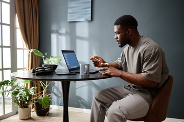 Young adult Black man sitting at round table eating meal while working on laptop near window, appearing focused and engaged with digital device during house arrest situation
