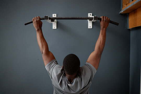 Black young adult man exercising by gripping pull up bar mounted on wall, viewed from behind, demonstrating strength training during house arrest in indoor setting
