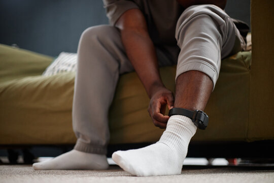 Black man sitting on sofa adjusting electronic ankle monitor on leg, wearing casual clothing and socks, concept of house arrest or home confinement, partial body visible