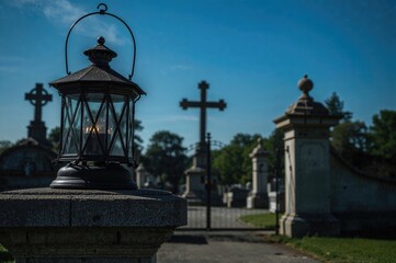 Fototapeta premium A glowing lantern and three memorial candles rest on a stone base with tombstones behind and the entrance gate of the burial ground under a clear blue sky