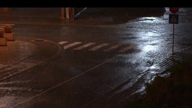 Pedestrian crosswalk and wet road during heavy rain. View of a pedestrian crosswalk and wet asphalt road during heavy rain. Street reflections from lamps create a moody urban scene.