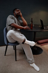 Black young adult man sitting at table wearing electronic ankle monitor, resting head on hand,...