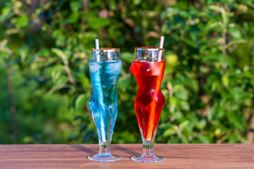 Two artistic glasses shaped like a nude woman and man filled with red and blue alcoholic cocktails on table with lush green garden background, closeup. Playful summer vibe for creative drink concepts