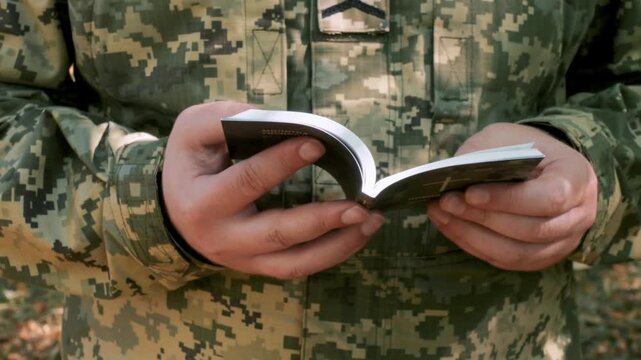 Military man in camouflage uniform holding and reading a small Bible outdoors, concept of faith, hope and spirituality in army life