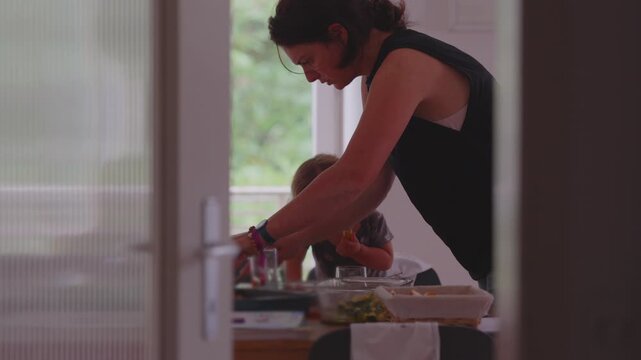 Mother serving food to young toddler during family meal, leaning over dining table with homecooked vegetables and fries, authentic moment of warmth and togetherness